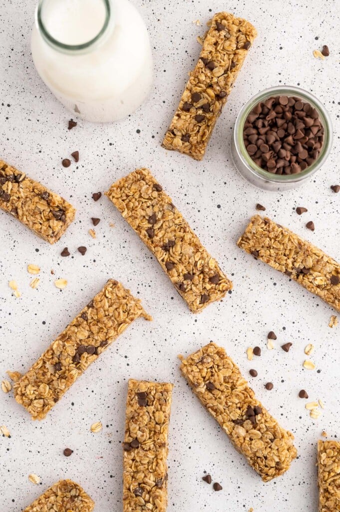 Homemade Healthy Granola Bars scattered on a grey counter top with mini chocolate chips and a bottle of milk on the side
