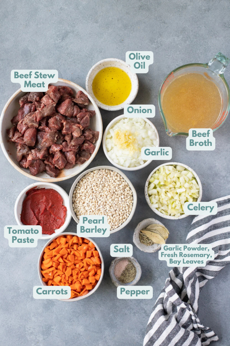 Ingredients for beef barley soup in white bowls: stew meat, olive oil, salt, pepper, garlic powder, rosemary, bay leaves, beef broth, pearl barley, veggies, tomato paste