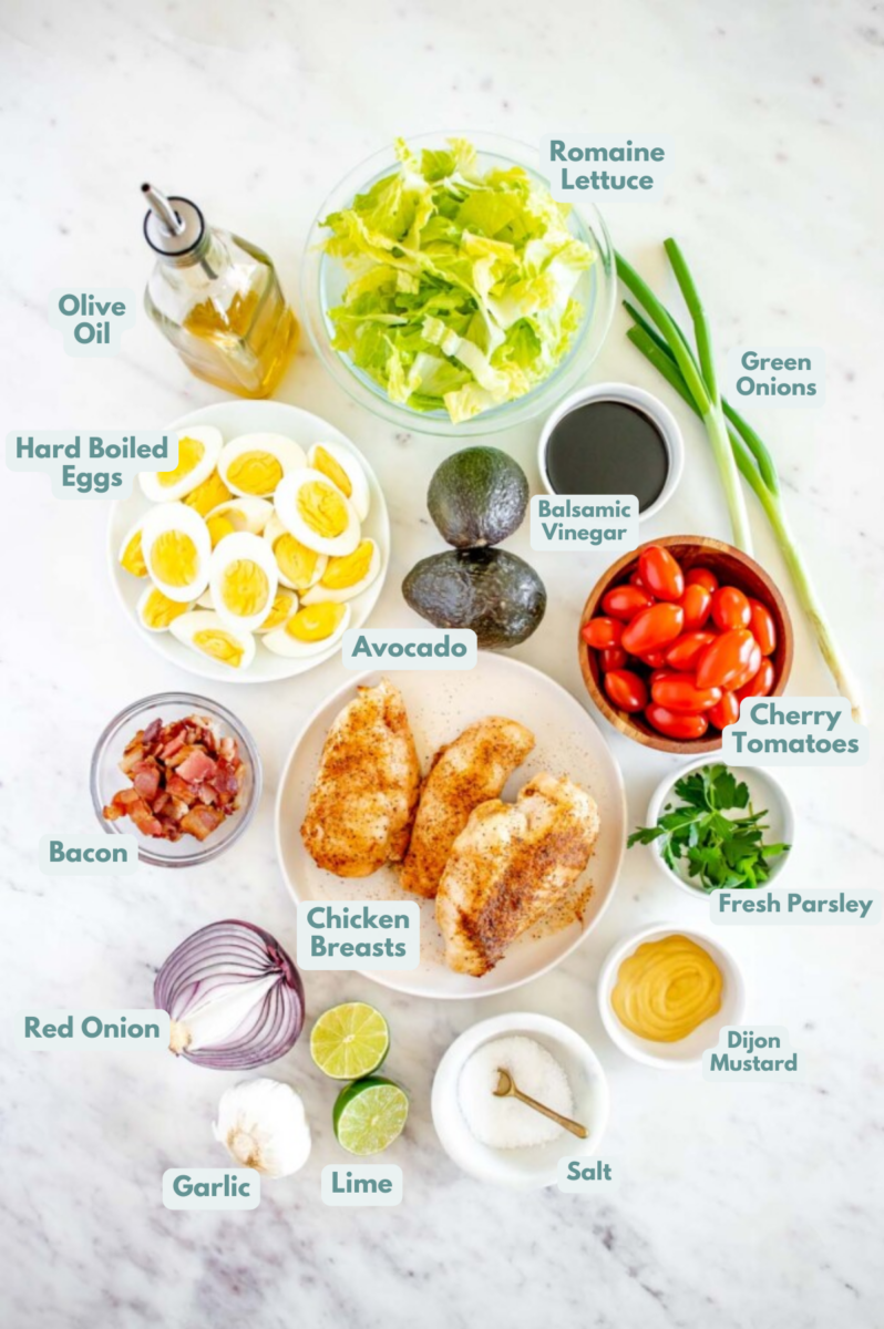 Ingredients for chicken cobb salad in small bowls: romaine lettuce, bacon, hard boiled eggs, avocado, cherry tomatoes, red onion, cooked chicken breasts, fresh parsley, green onions, balsamic vinegar, dijon mustard, garlic, olive oil, lime juice, salt, pepper