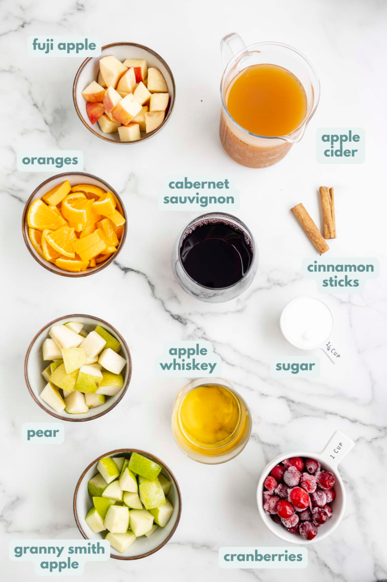Ingredients for Red Wine Sangria in small bowls: water, granulated sugar, cinnamon sticks, cabernet sauvignon, apple cider, apple whiskey, cranberries, pear, granny smith apple, fuji apple, orange