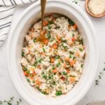 An overhead shot of crockpot chicken and rice in a white slow cooker base with a gold spoon in it with some herbs on the side on a white background.