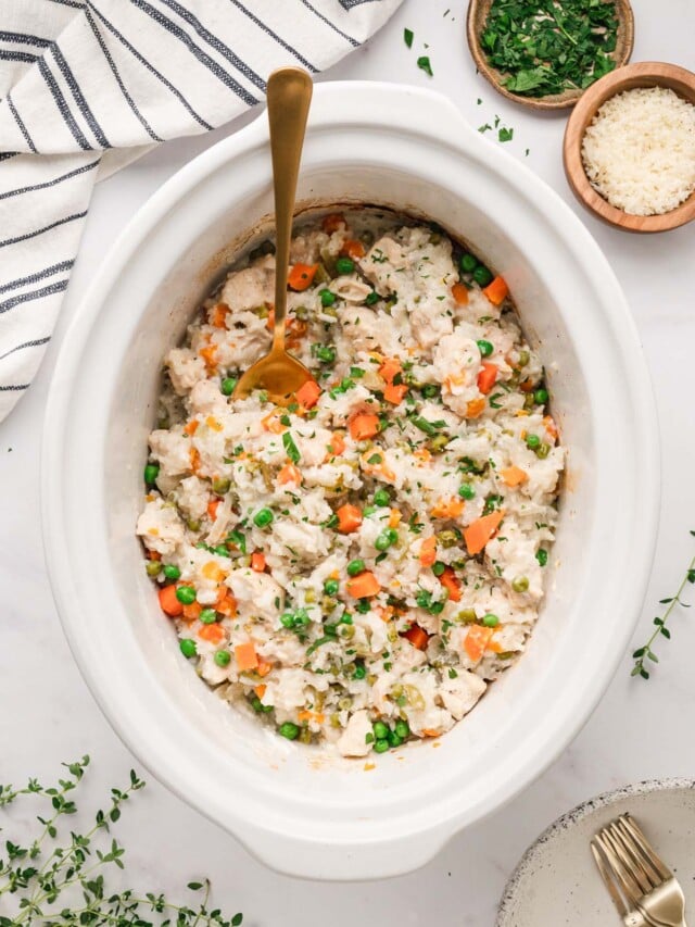 An overhead shot of crockpot chicken and rice in a white slow cooker base with a gold spoon in it with some herbs on the side on a white background.