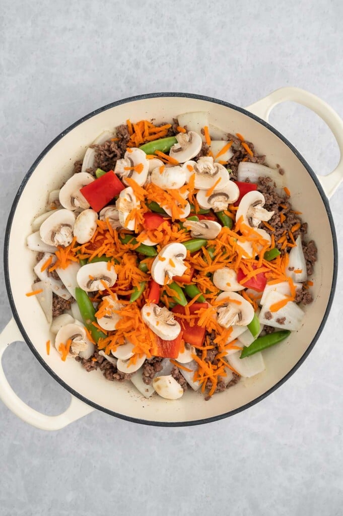 Raw stir fry vegetables piled on cooked ground beef, onions, and garlic in a dutch oven.
