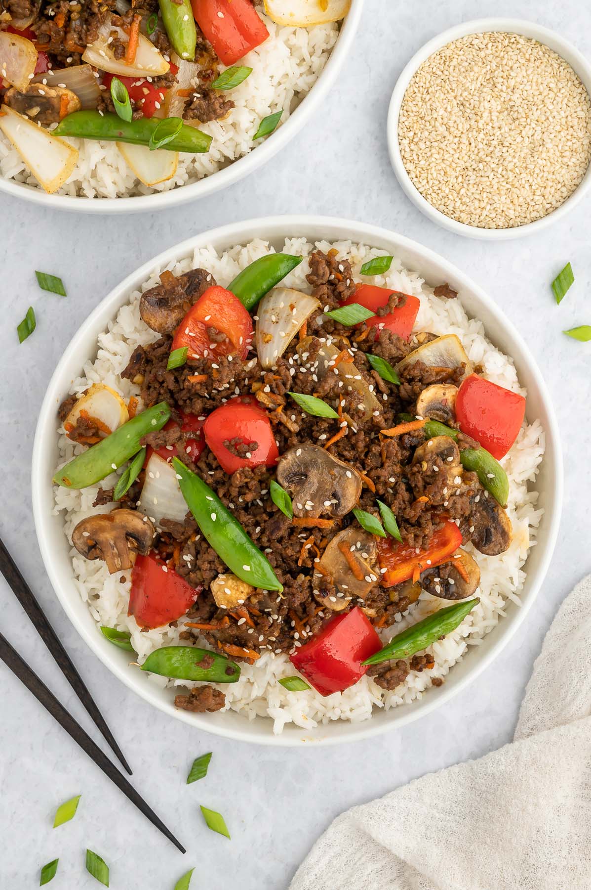 Plated ground beef stir fry on a bed of rice next to a bowl of sesame seeds.