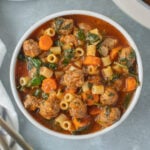 Meatball Soup in a white bowl with a pot and bowl of soup on the side on a grey background