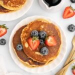 Protein Pancakes stacked on a white plate with strawberries and blueberries on top with another plate of pancakes, fruit and a small white pitcher of maple syrup on the side.