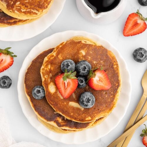 Protein Pancakes stacked on a white plate with strawberries and blueberries on top with another plate of pancakes, fruit and a small white pitcher of maple syrup on the side.