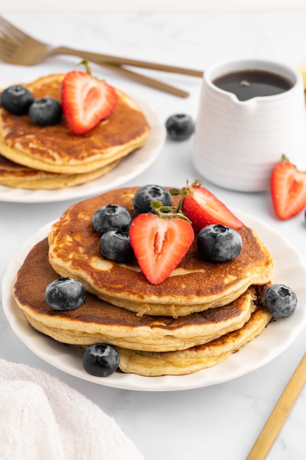 Stack of three high protein pancakes topped with strawberries and blueberries on a white plate next to a small white pitcher of maple syrup.