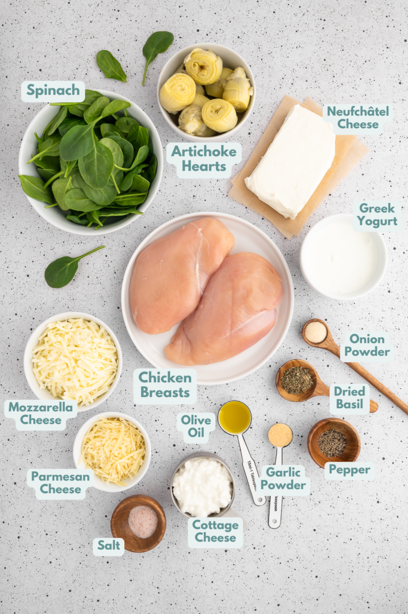 Ingredients for spinach artichoke chicken in small bowls on a grey counter: chicken breast, olive oil, neufchâtel cheese, greek yogurt, cottage cheese, mozzarella cheese, shredded parmesan, spices, artichoke hearts, spinach
