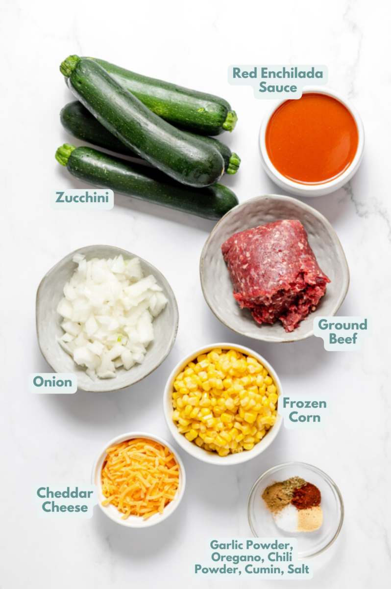 Ingredients for Stuffed Zucchini in small bowls on a white counter: olive oil, ground beef, onion, frozen corn, zucchini, spices, cheddar cheese, red enchilada sauce
