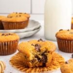 A Healthy Chocolate Chip Muffin with a bite taken out of it sitting on it's cupcake liner with more cupcakes scattered around on a white counter.
