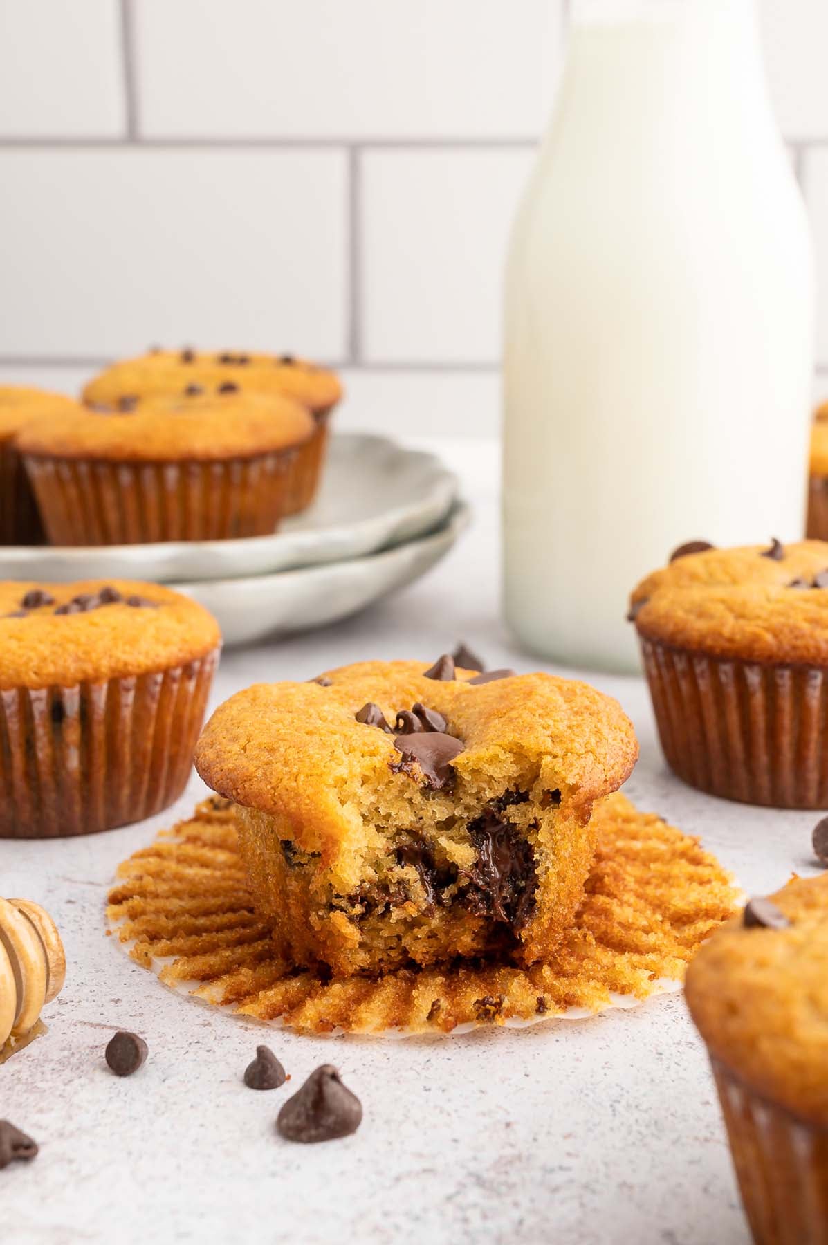 A healthy chocolate chip muffin with a bite taken out of it sitting on its paper wrapper with more muffins scattered around on a white counter.