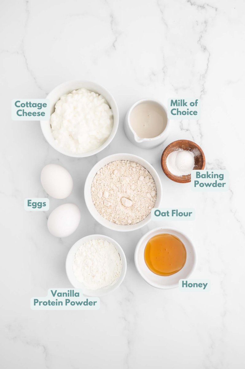 Ingredients for Protein powder pancakes in small bowls on a white background: Oat flour, vanilla protein powder, honey, eggs, cottage cheese, milk of choice, baking powder
