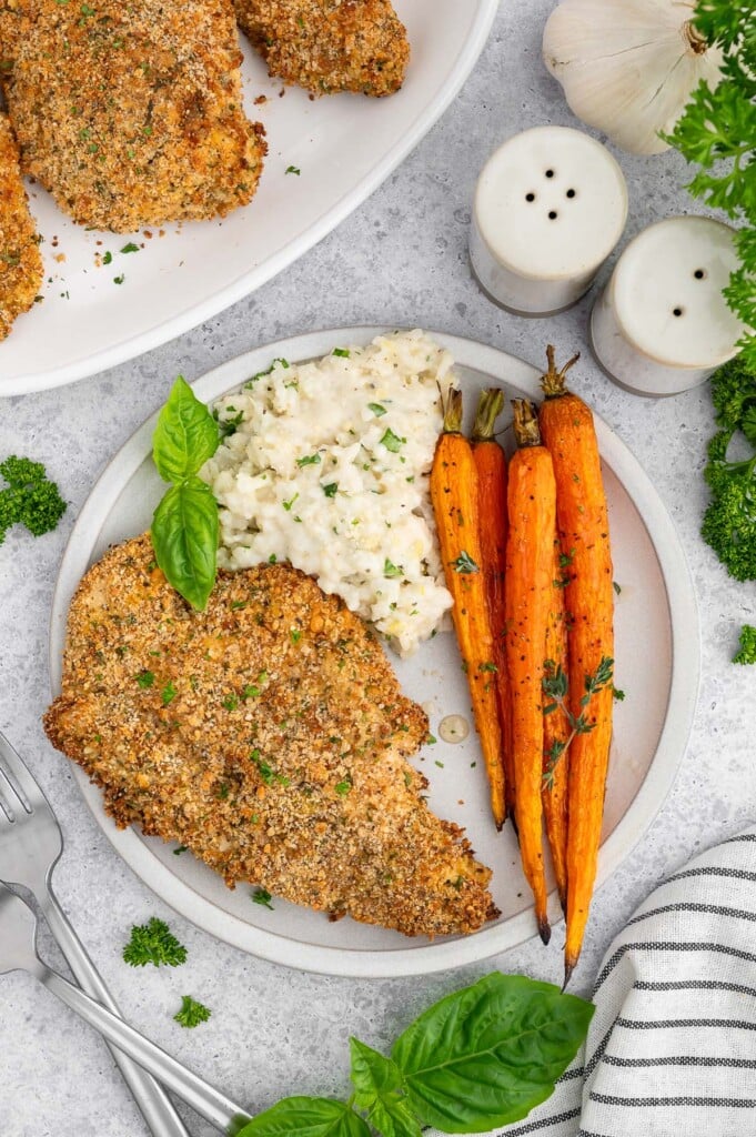 A plate with a baked chicken cutlet, lemon risotto, and roasted carrots with a plate of more cutlets on the side.