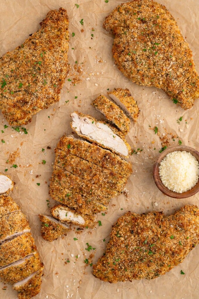 5 Baked Chicken Cutlets on a piece of parchment paper. Two of the cutlets are sliced.