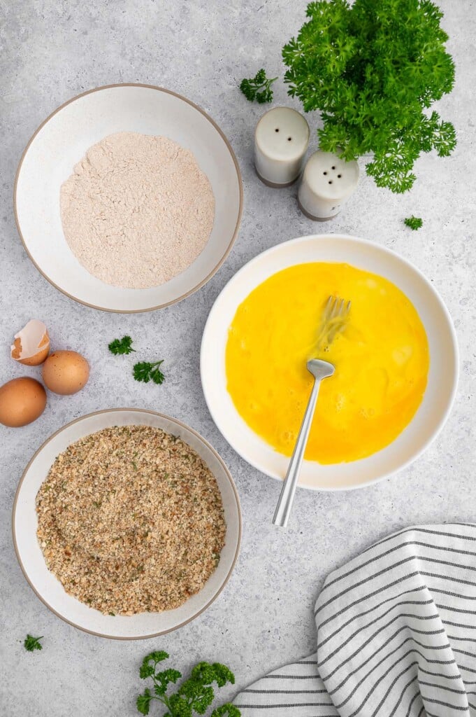 Three bowls on a white background: one has whole wheat flour, one has breadcrumbs with spices and the third has scrambled eggs with a fork in it.