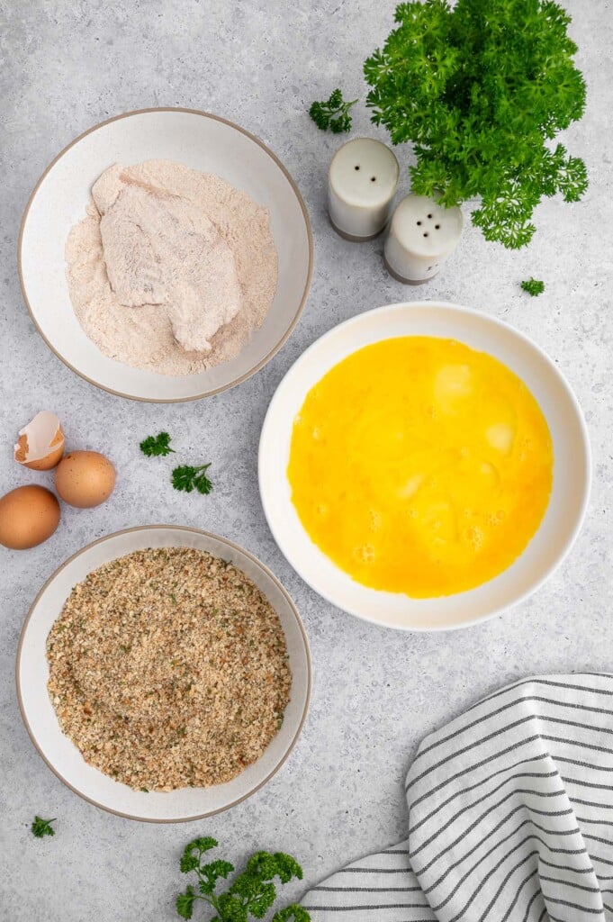 A chicken cutlet in a bowl of whole wheat flour with two bowls of eggs and seasoned breadcrumbs on the side.