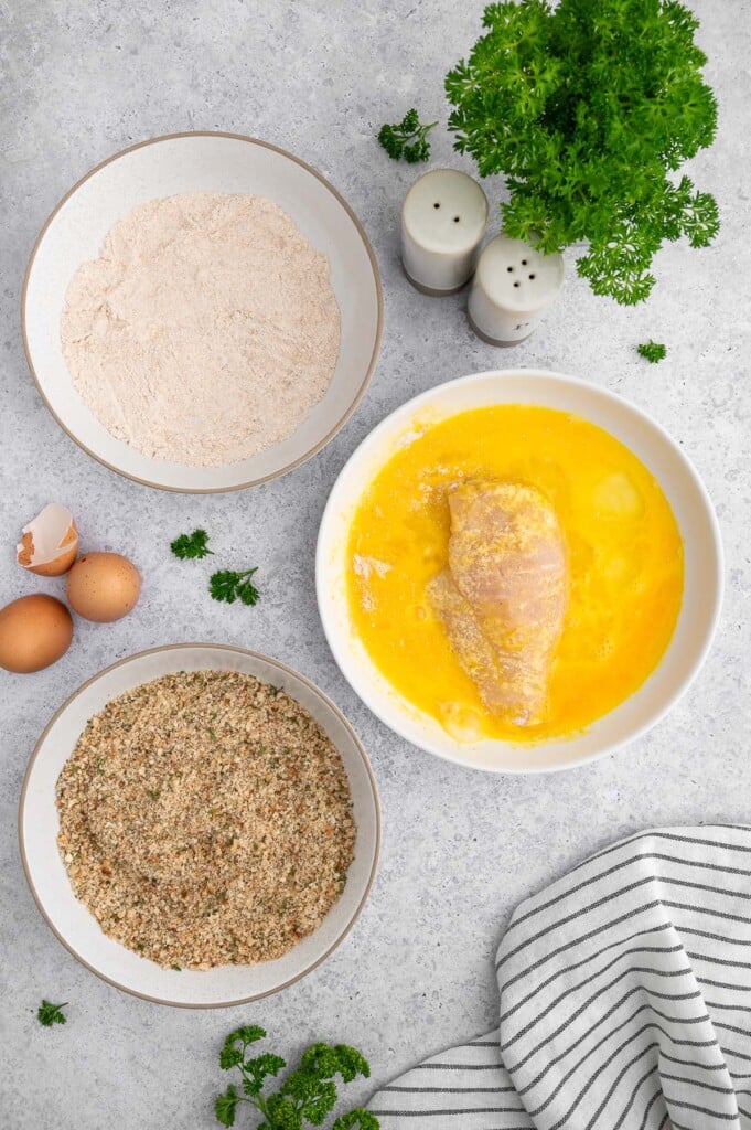 A chicken cutlet that has been dipped in flour and eggs in the bowl of eggs with two bowls of whole wheat flour and seasoned breadcrumbs on the side.