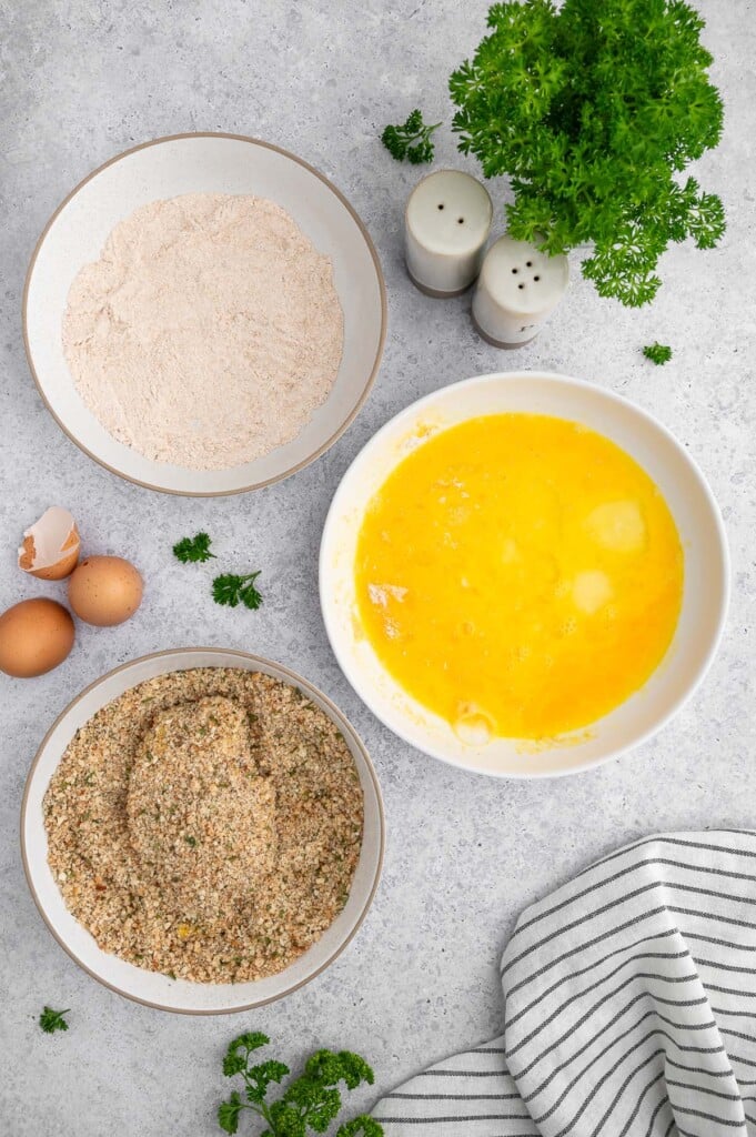 A fully breaded chicken cutlet in a bowl of seasoned breadcrumbs with a bowl of whole wheat flour and eggs on the side.