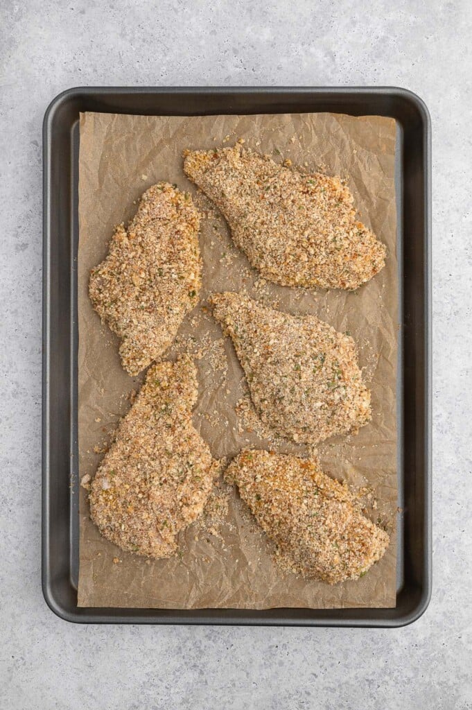 Uncooked breaded chicken cutlets on a parchment lined baking sheet.