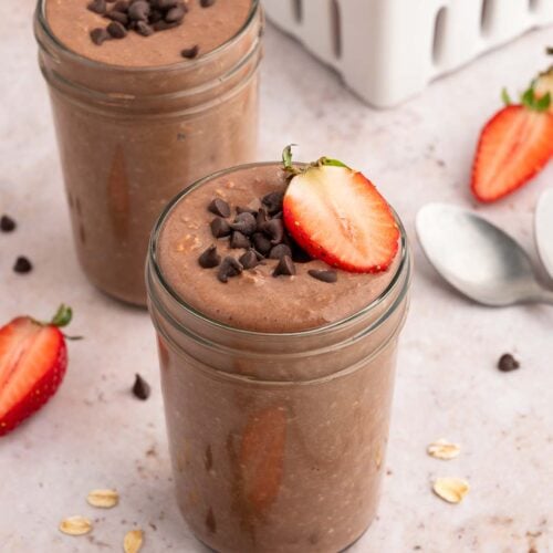 Two mason jars with chocolate overnight oats topped with mini chocolate chips and sliced strawberries.