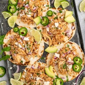 5 Crockpot Chicken Tacos on a baking sheet garnished with jalapeños, lime slices and avocado pieces.
