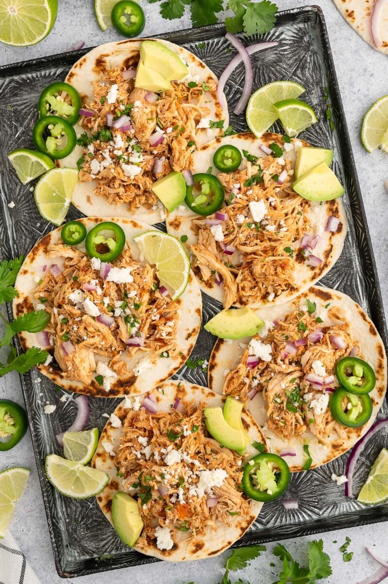 5 Crockpot Chicken Tacos on a baking sheet garnished with jalapeรฑos, lime slices and avocado pieces.