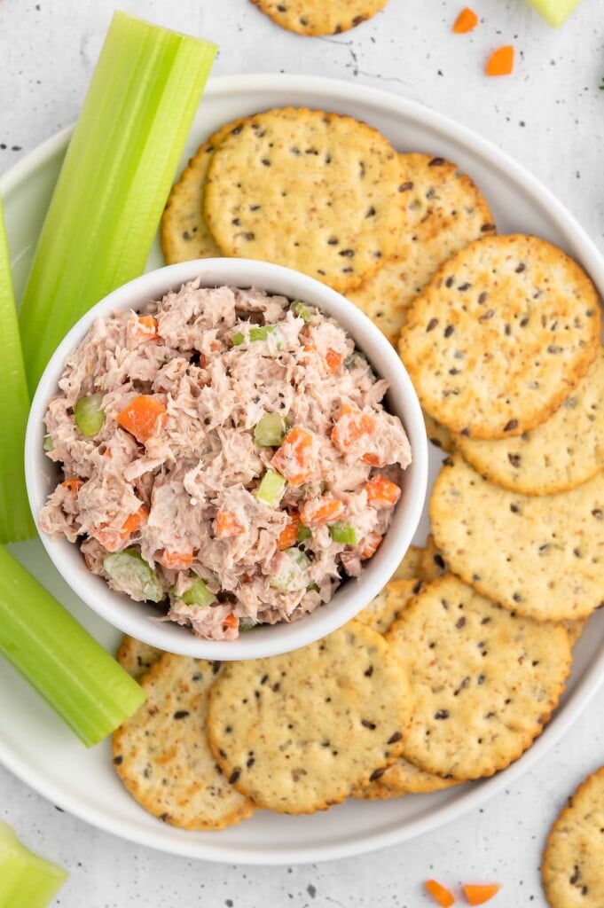 A bowl of healthy tuna salad on a plate with crackers and celery sticks on the side.