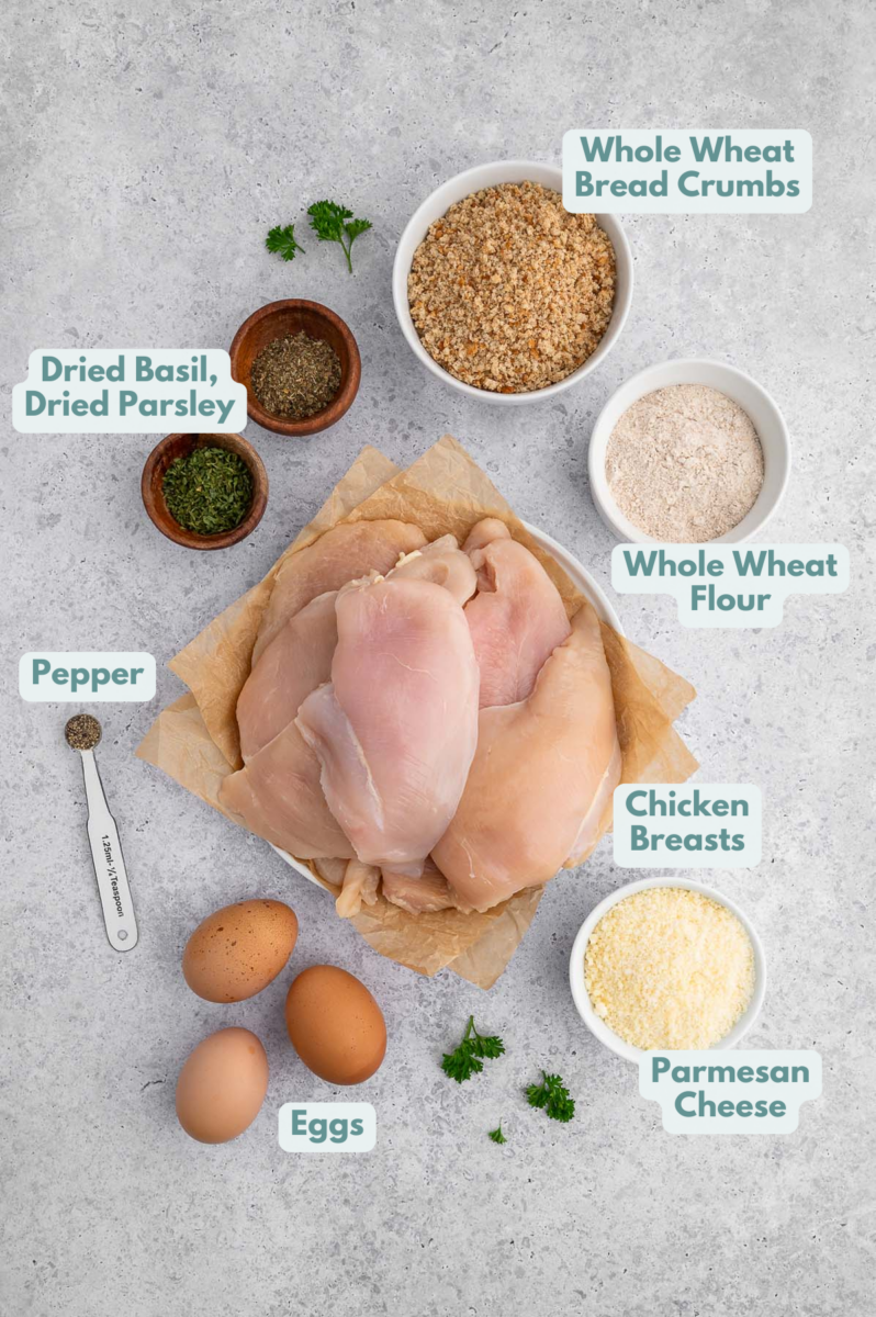 Ingredients for Baked Chicken Cutlets in small bowls: chicken breasts, whole wheat flour, egg, whole wheat breadcrumbs, parmesan cheese, pepper, dried basil, dried parsley