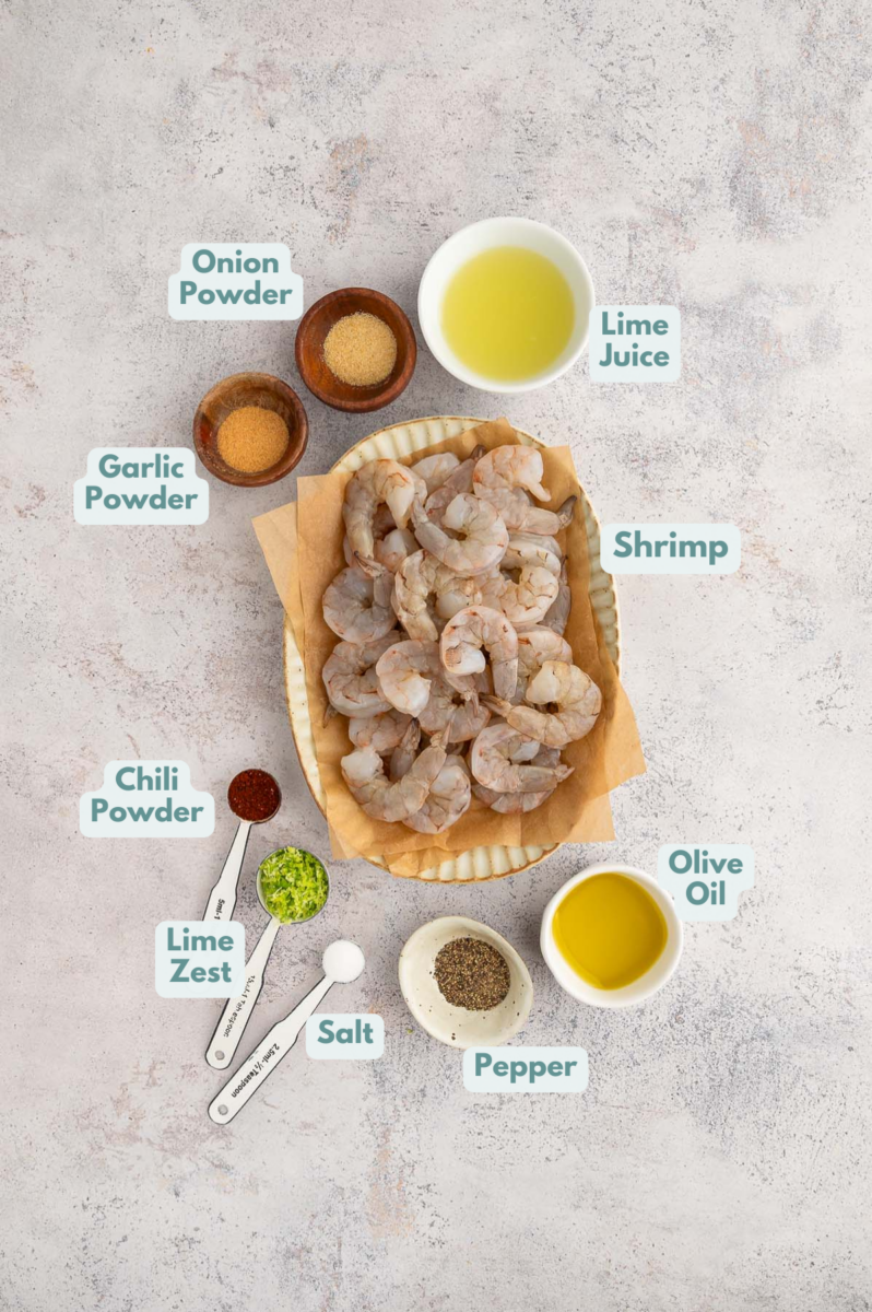 Ingredients to make chili lime shrimp in white bowls on a grey background: shrimp, fresh lime juice, lime zest, spices including chili powder, olive oil