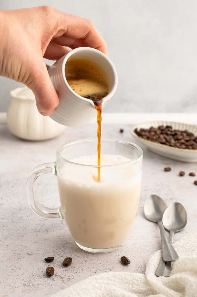 A hand pouring espresso into a clear mug with milk to make a healthy vanilla latte.