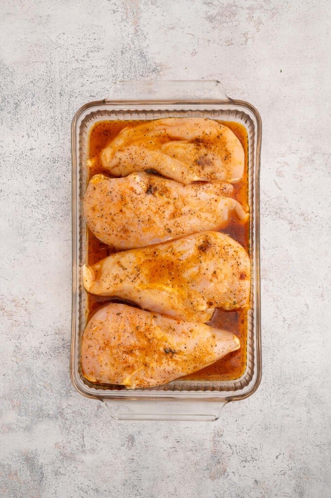 Four boneless skinless chicken breasts in a glass baking dish of marinade.