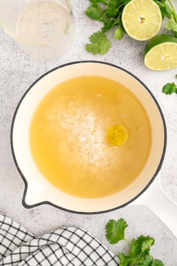 Uncooked chicken broth, white rice, and spices in a pot with limes and cilantro on the side. 