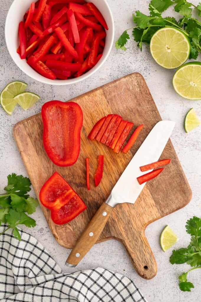 A knife on a cutting board with partially sliced red bell peppers and a bowl of the sliced peppers on the side. 