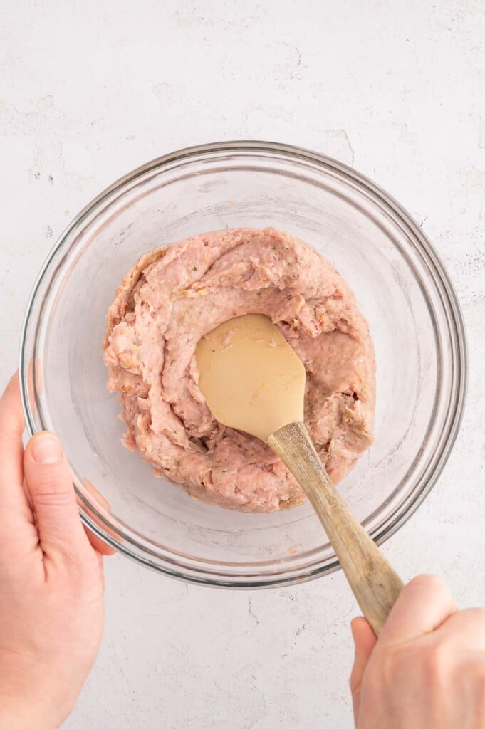 Ground turkey, maple syrup, red wine vinegar, and spices in a clear mixing bowl being mixed with a rubber spatula.