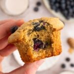 A hand holding up a blueberry protein muffin with a bite taken out of it.