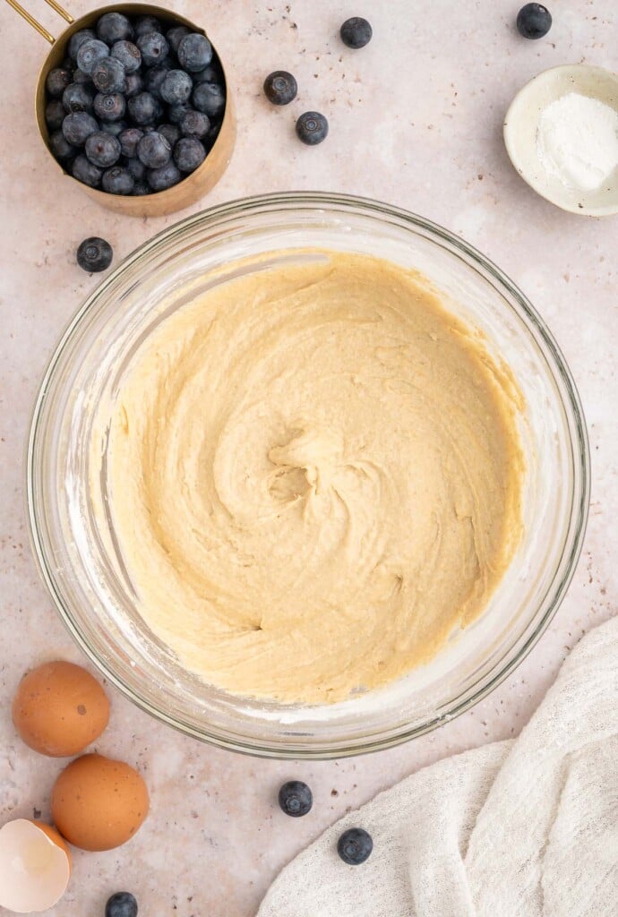A clear bowl with muffin batter mixed together with blueberries on the side.