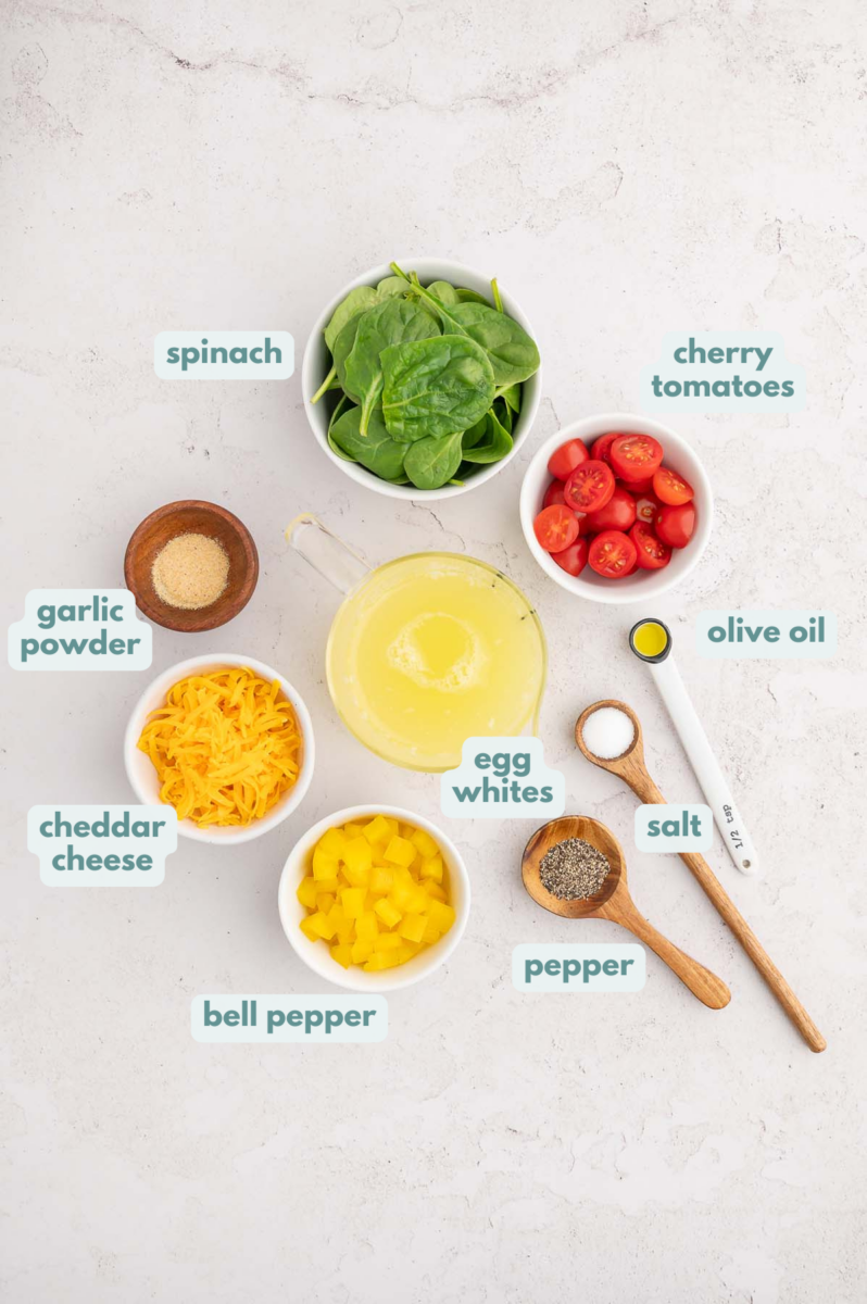 Ingredients for egg white frittata in small bowls: olive oil, bell pepper, spinach, cherry tomatoes, egg whites, shredded cheddar cheese, black pepper, garlic powder, salt