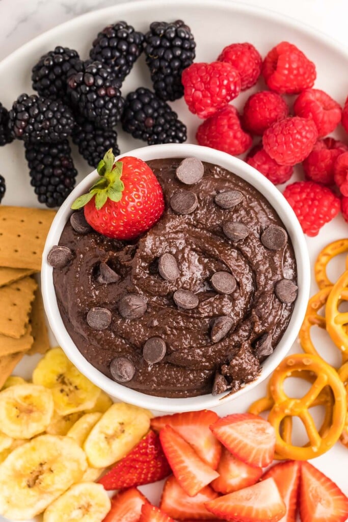 Chocolate hummus in a small white bowl topped with chocolate chips and a strawberry surrounded by blackberries, raspberries, strawberries, banana slices, pretzels, and graham crackers. 
