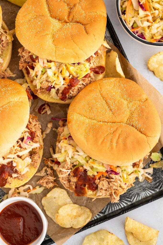 An overhead shot of 3 bbq chicken and coleslaw sandwiches on a platter with a small bowl of vinegar coleslaw, bbq sauce, and chips on the side.