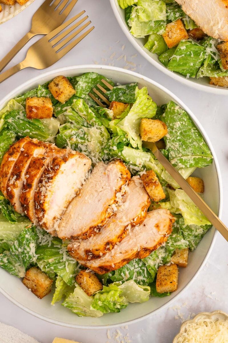A sliced grilled chicken caesar salad in a white bowl topped with homemade croutons and a fork in the bowl.