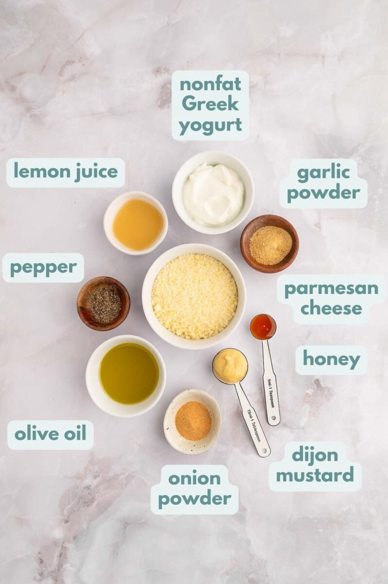 Ingredients for caesar salad dressing in small bowls, labeled: nonfat Greek yogurt, garlic powder, parmesan cheese, honey, dijon mustard, onion powder, olive oil, pepper, lemon juice
