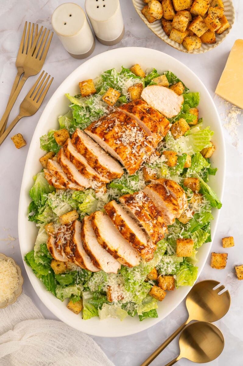 A white platter of a caesar salad with two sliced grilled chicken breasts on top. 