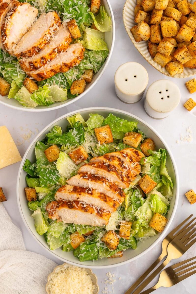 An overhead shot of a chicken caesar salad topped with homemade croutons in a bowl with another salad and a platter of croutons on the side.