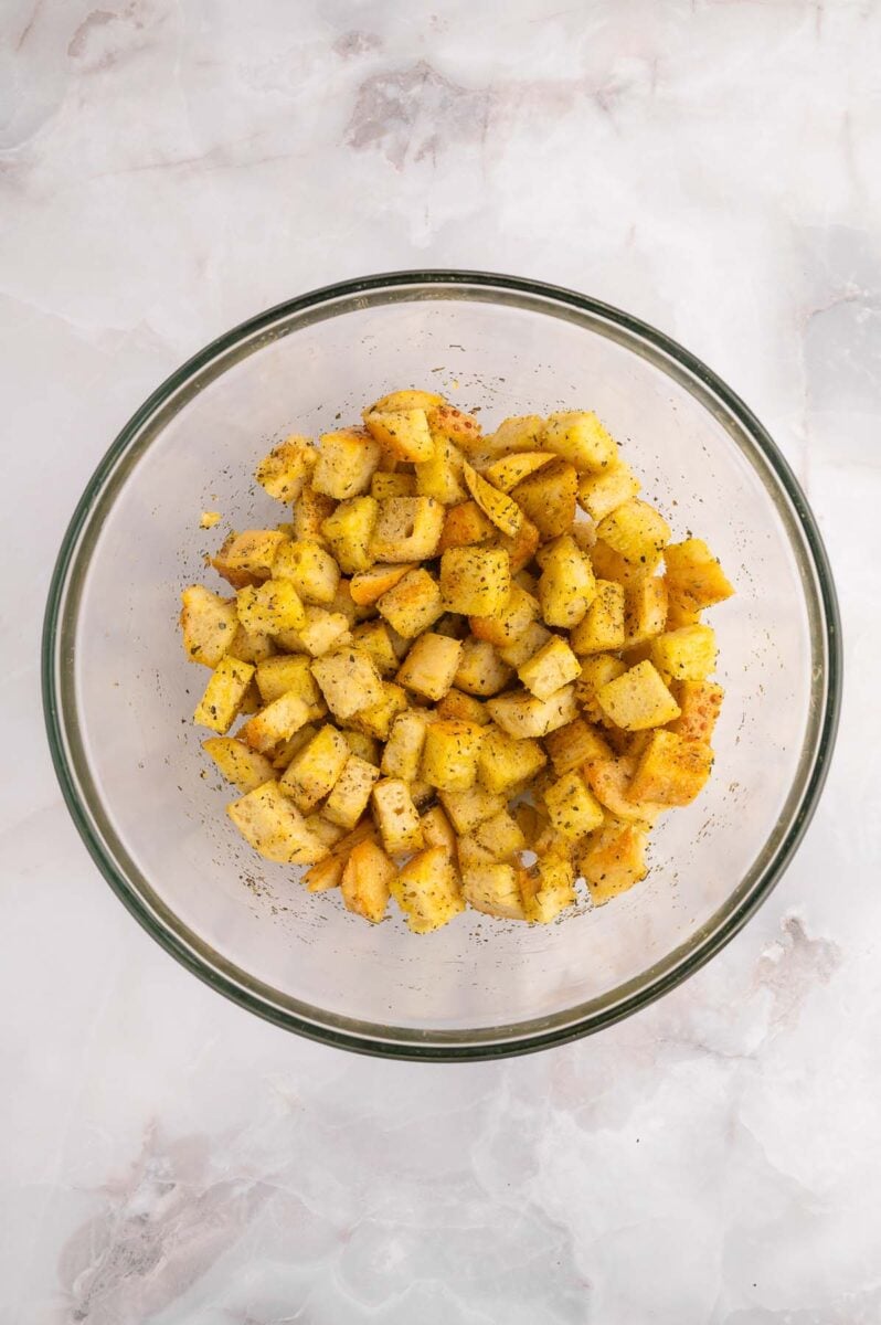 Uncooked cubed bread tossed in olive oil and spices in a clear bowl.