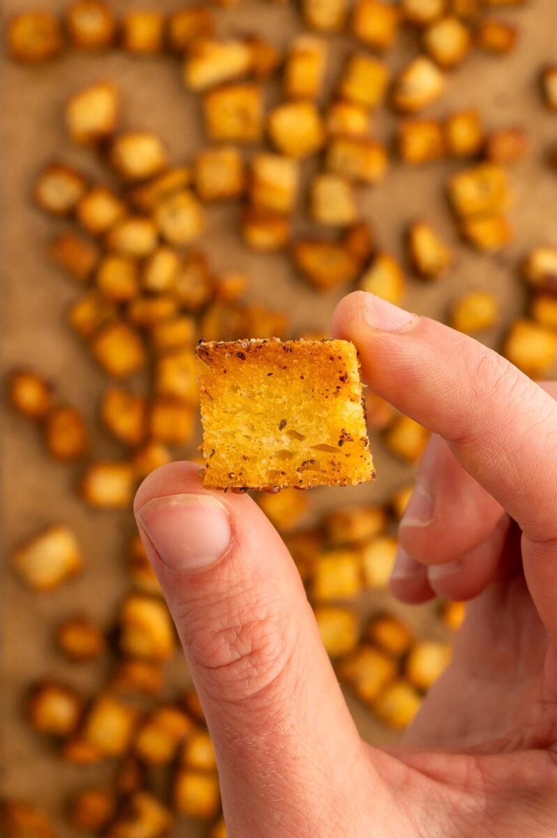 A hand holding up a crouton with a parchment lined baking sheet of croutons in the background.