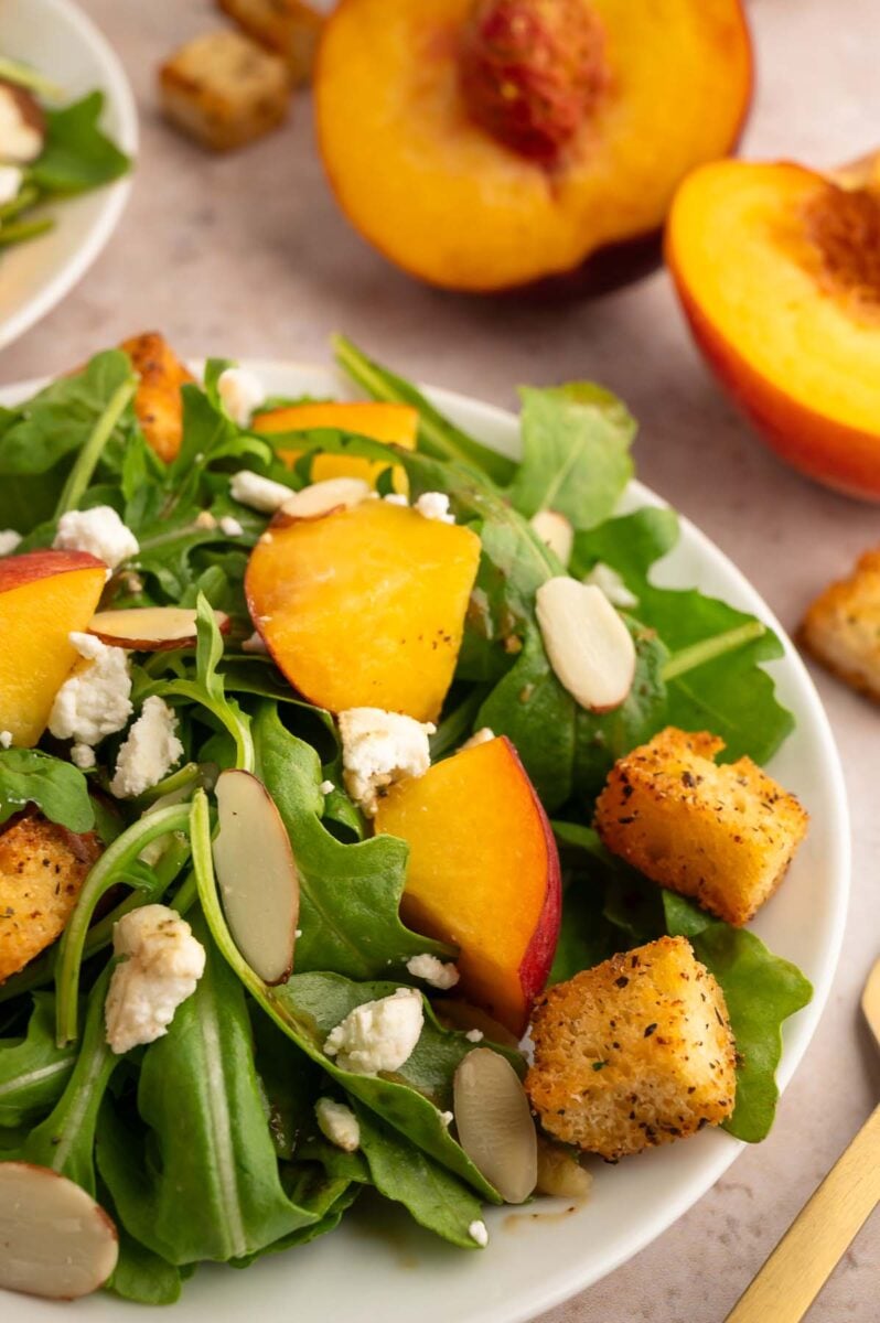 A peach feta salad topped with homemade croutons with fresh peaches in the background.