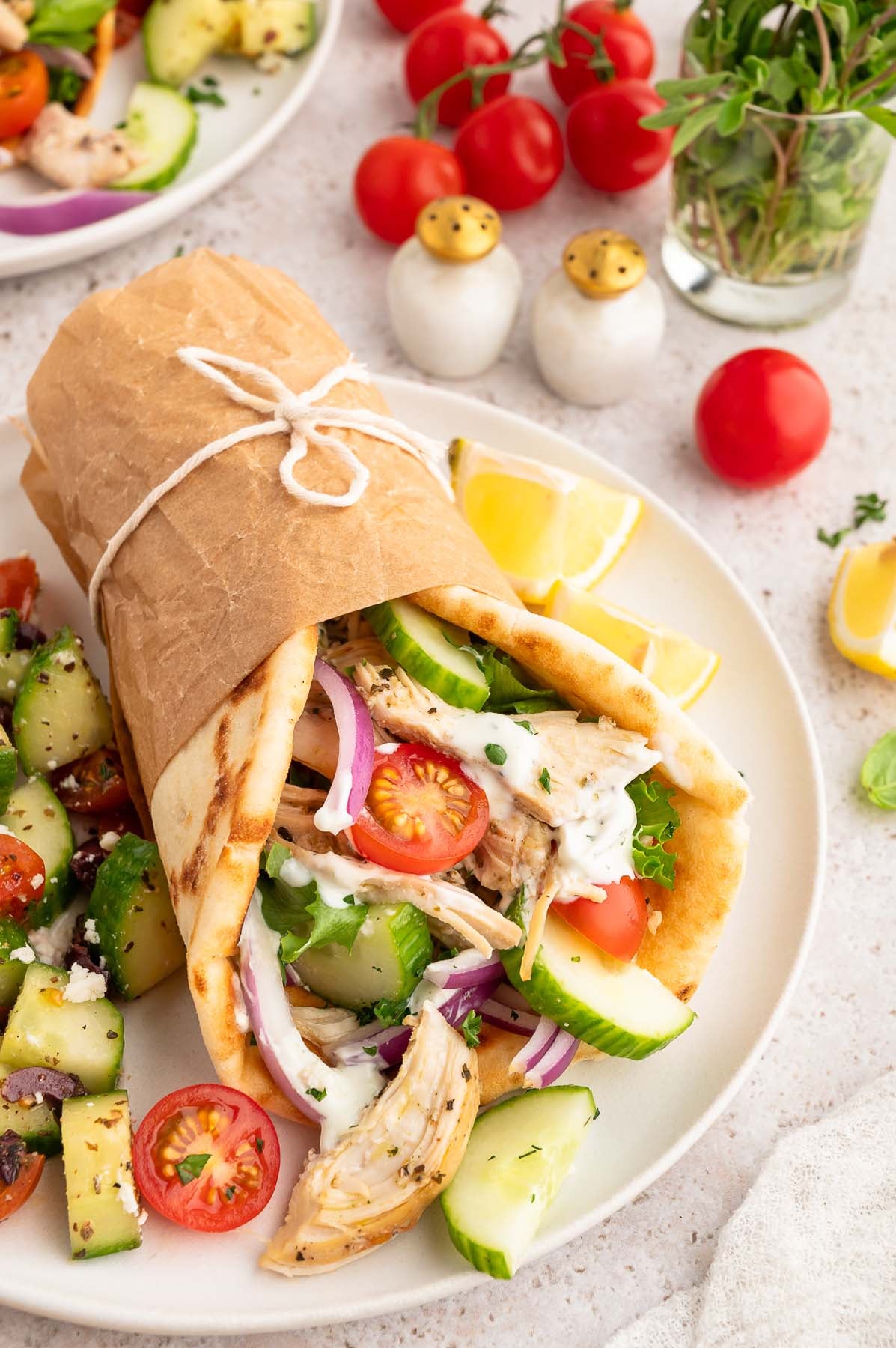 Greek shredded chicken wrapped in a pita with cherry tomatoes, red onion slices, and cucumber slices. Sandwich is wrapped in paper and secured with twine.