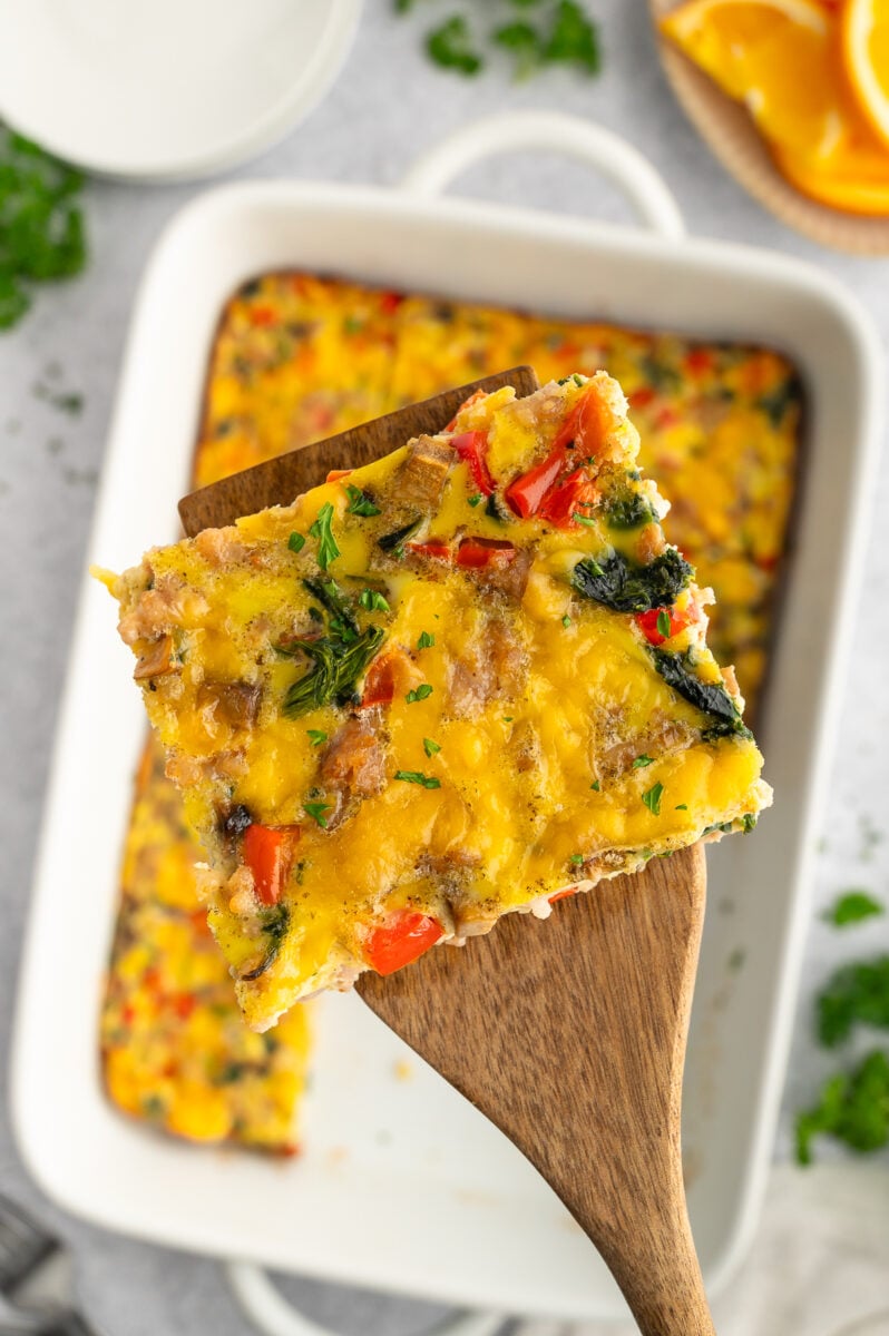 A piece of sausage egg bake lifted up on a wooden spoon with the casserole dish in the background.
