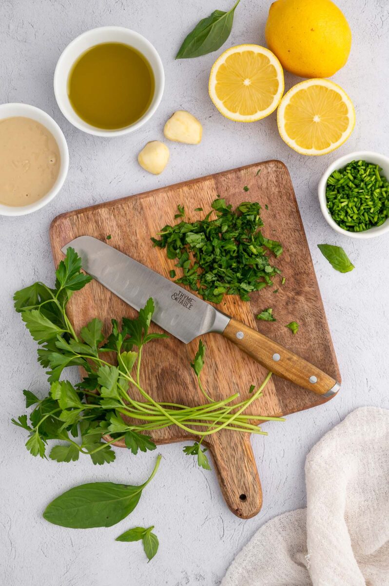 Fresh basil, parsley and chives chopped on a wooden cutting board with a knife on it and ingredients for tahini dressing on the side.
